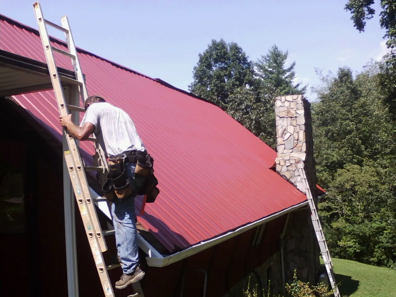 Roofer on a ladder beside a red metal roof and stone chimney — Soffit & Fascia Repair by Roofing Brothers in Algona